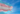 Ao fundo, o céu azul, entrando na foto pelo lado esquerdo, uma bandeira representa a luta trans, com listras na seguinte padronagem: azul, rosa, branco, rosa e azul; exemplifica a visibilidade trans nas empresas.